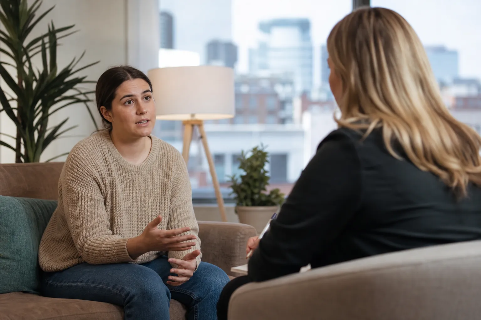 Two Women Talking in a Bright Office A woman in a beige sweater gestures while talking to a seated woman holding a pen in a bright office.