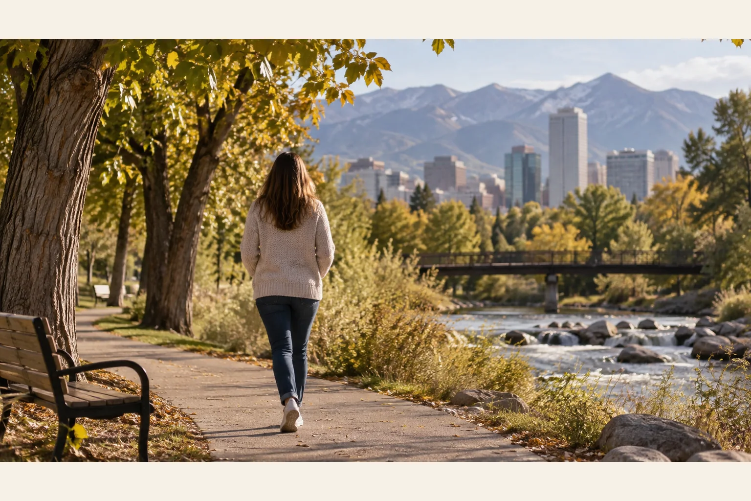 Autumn Riverside Walk With City and Mountain Views A woman walks along a tree-lined riverside path in autumn, with a bench, bridge, city skyline and mountains beyond.