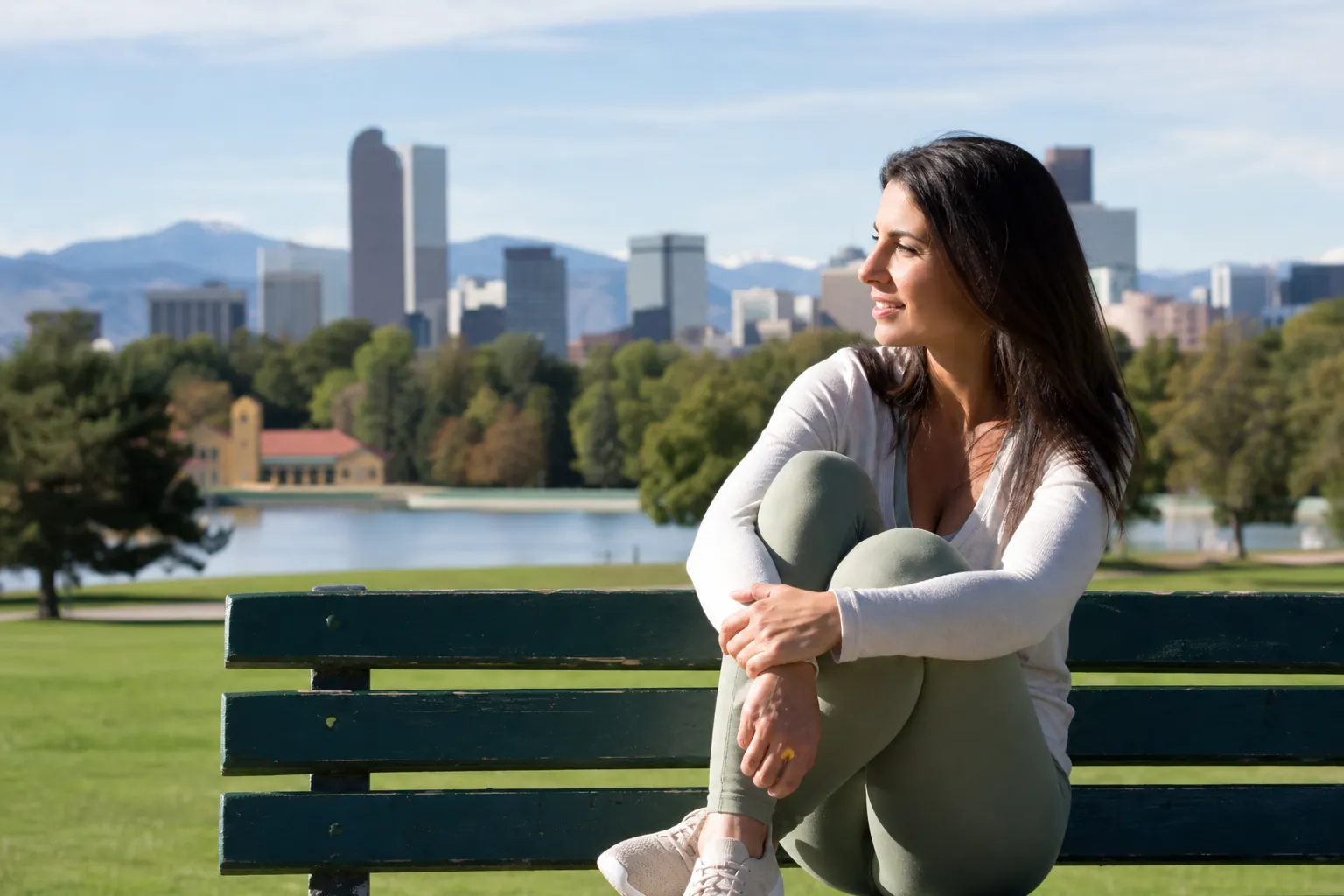 A woman sits with her knees drawn up on a green park bench beside a lake, with Denver's skyline and mountains behind her.