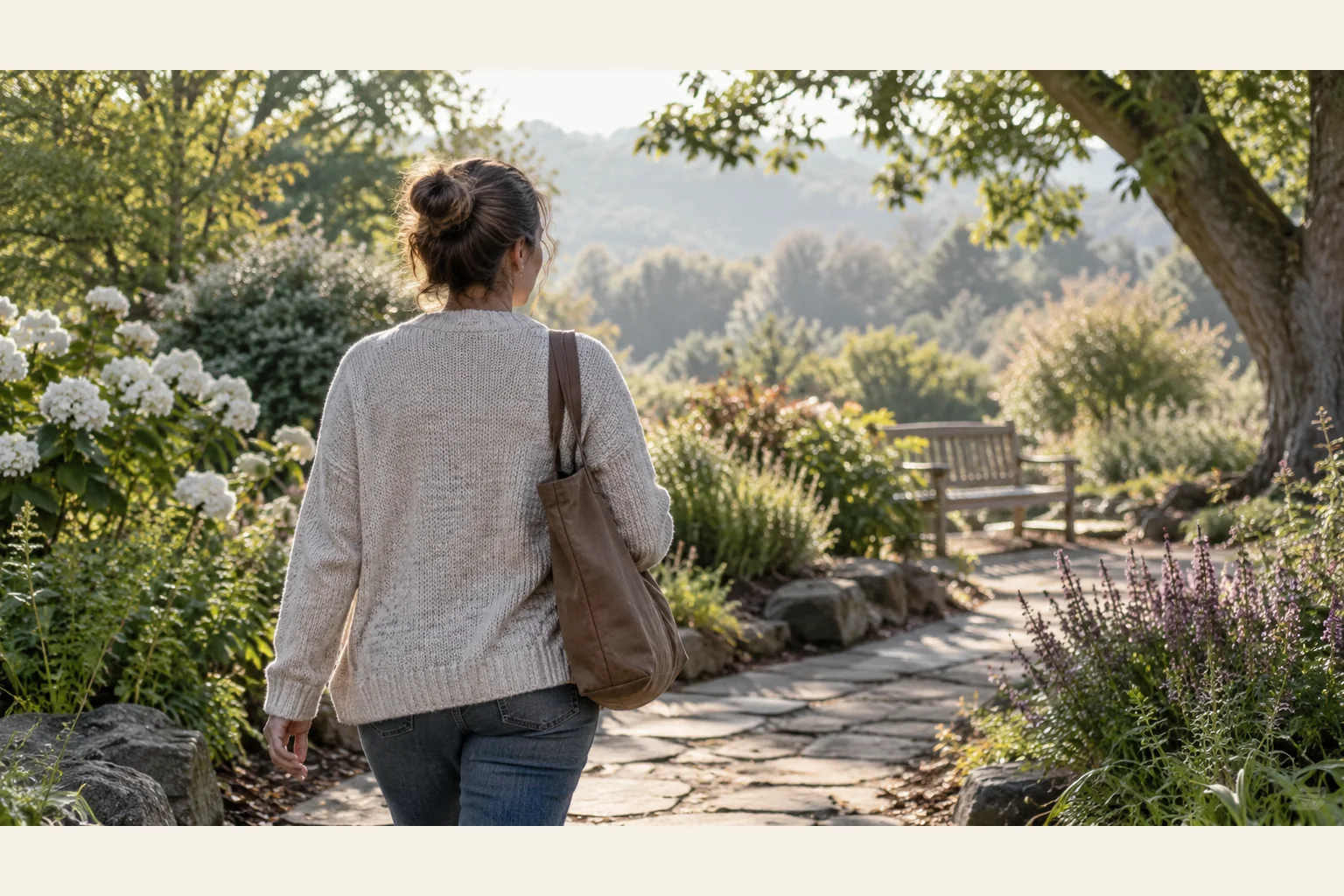 Woman Walking Along a Sunlit Garden Path A woman seen from behind carries a brown shoulder bag along a sunlit stone garden path toward a wooden bench.