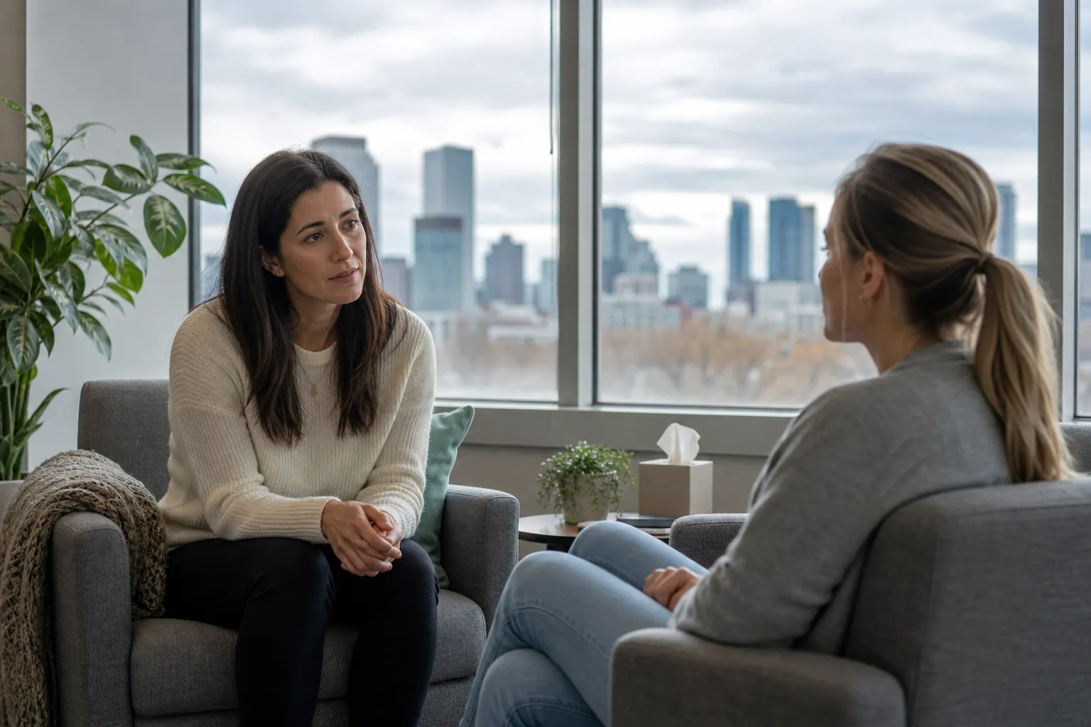 Two Women Talking in a Bright Office Two women sit facing each other in gray armchairs in a bright office with plants and large windows overlooking a city.