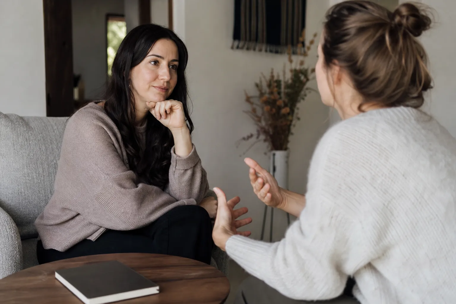 Illustrative scene: Two Women Seated in Conversation Illustrative scene: Two women sit facing each other in conversation, one listening with her hand under her chin while the other gestures.