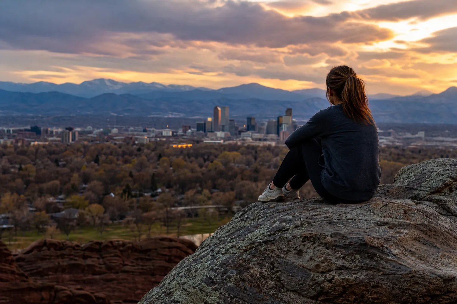 Woman Overlooking a City and Mountains at Sunset A woman sits with her knees drawn up on a rocky overlook, facing a city skyline and distant mountains at sunset.