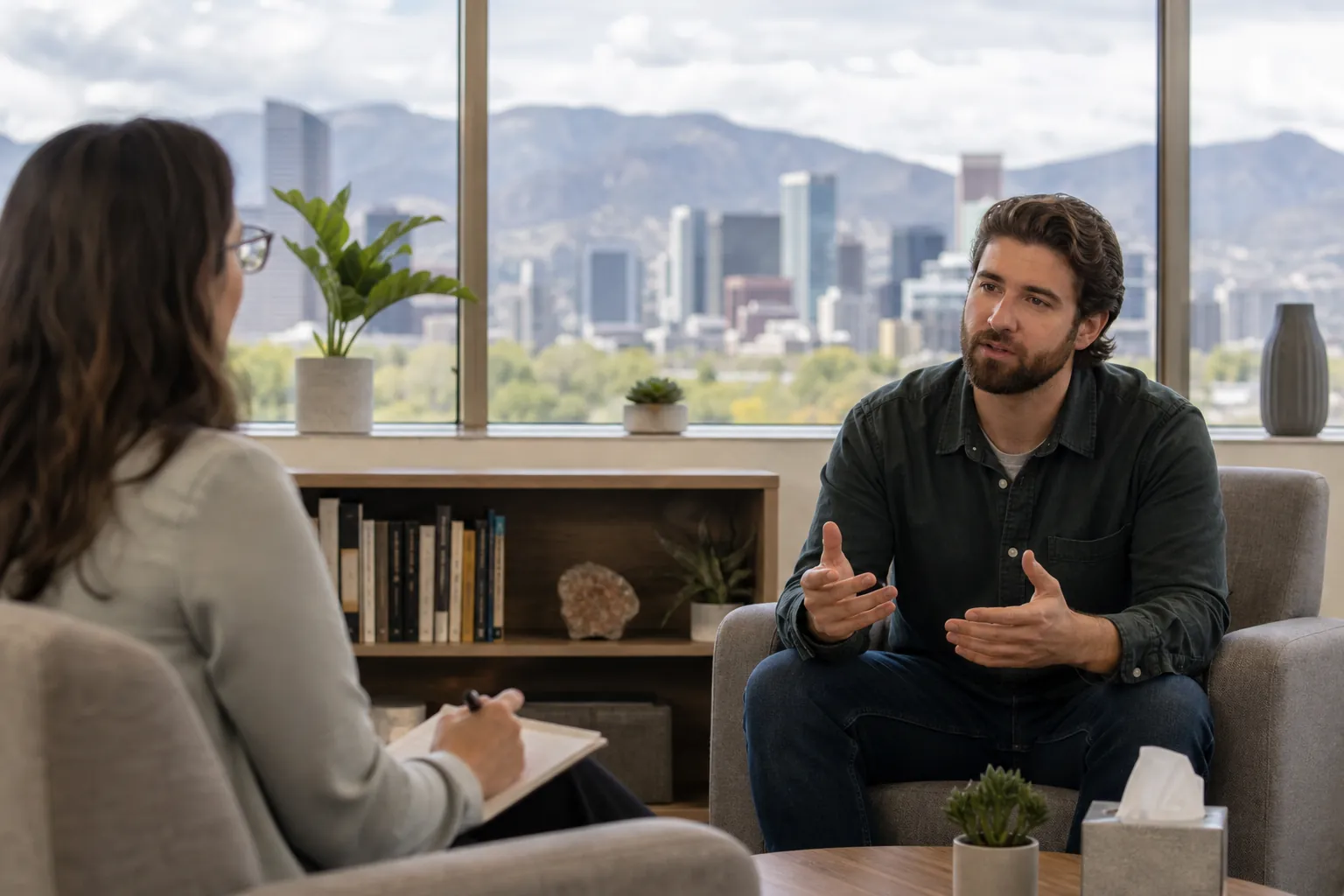 Individual Therapy Conversation in a Mountain View Office A man gestures while talking with a woman holding a notepad in an office overlooking a city skyline and mountains.