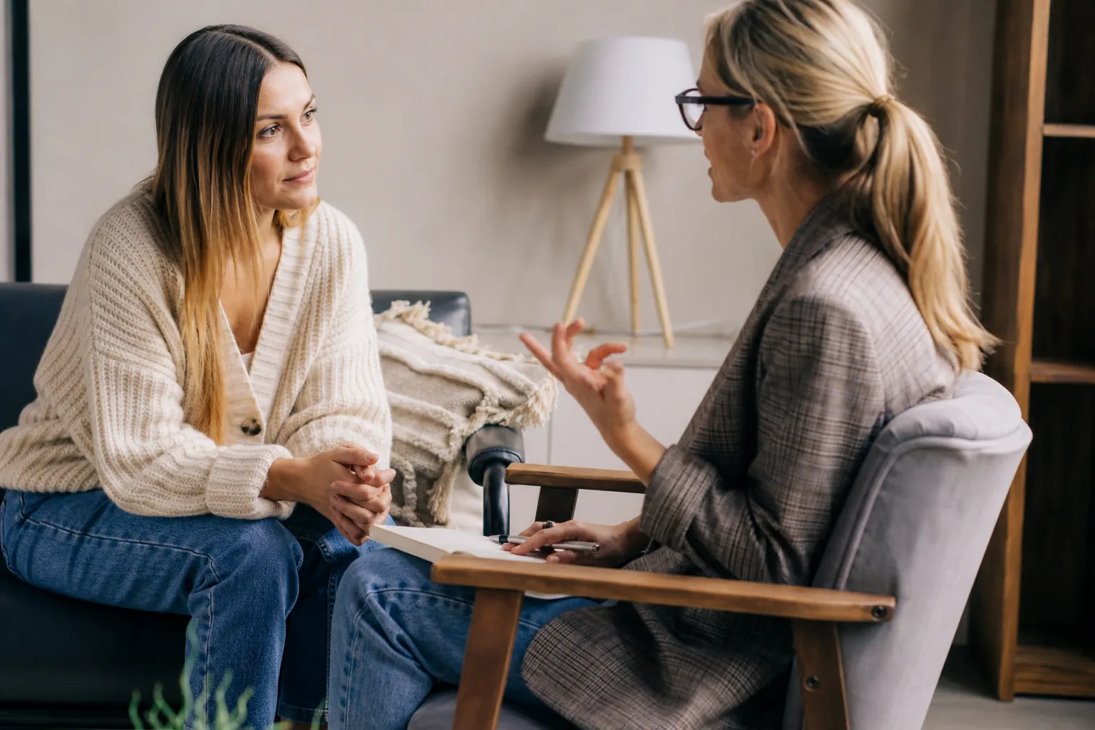 Two Women Talking in a Calm Counseling Setting Two women sit facing each other in a calm counseling setting, with one gesturing and holding a notebook on her lap.