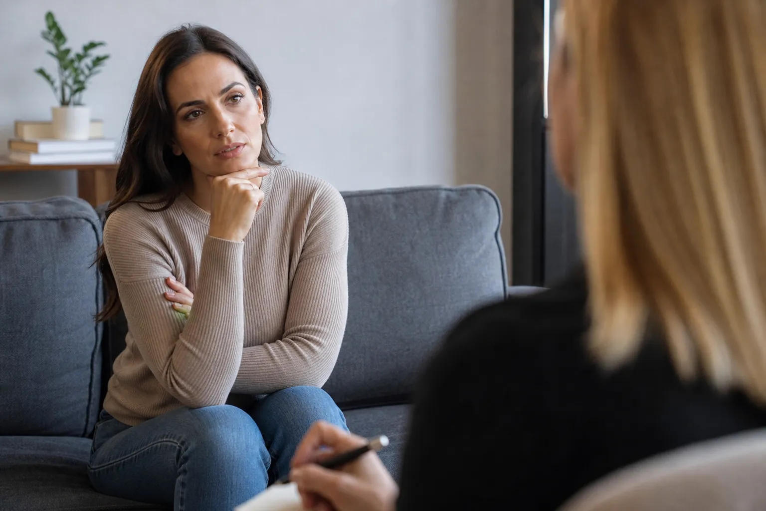 Thoughtful Client During a Therapy Session A thoughtful woman sits on a gray couch with her arms crossed, facing a blonde woman holding a pen during a therapy session.