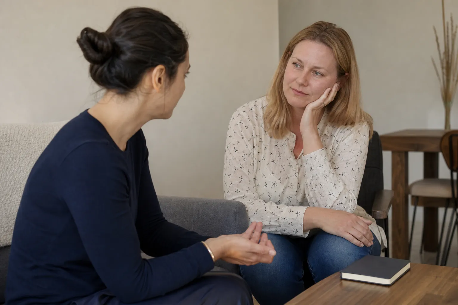 Two Women Seated in an Attentive Conversation Two women sit facing each other in conversation, one resting her cheek on her hand, with a closed notebook on a wooden table.