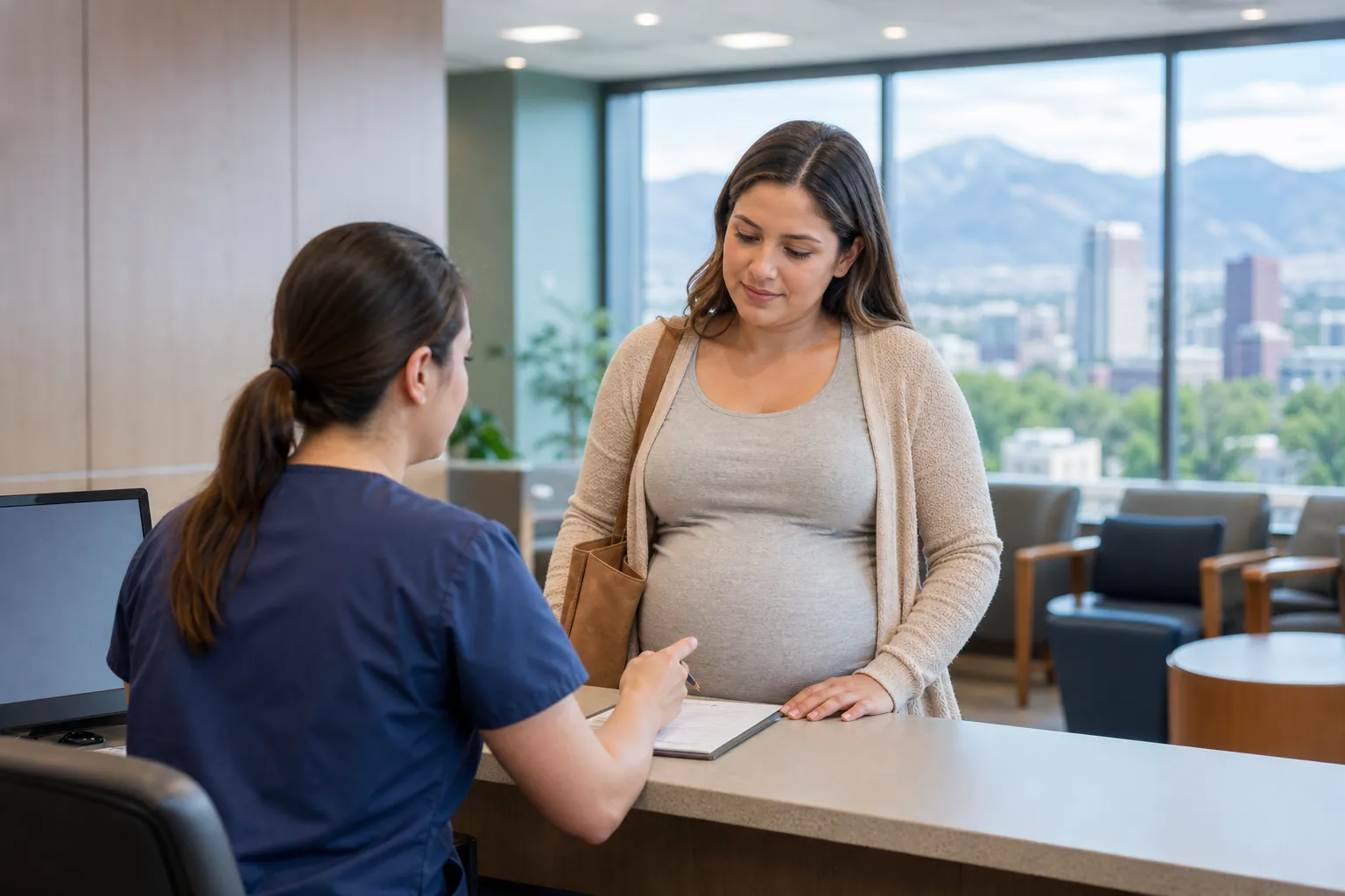 Pregnant Woman Speaking With Admissions Staff A pregnant woman speaks with a staff member in navy scrubs at a reception counter with paperwork and mountain views behind them.