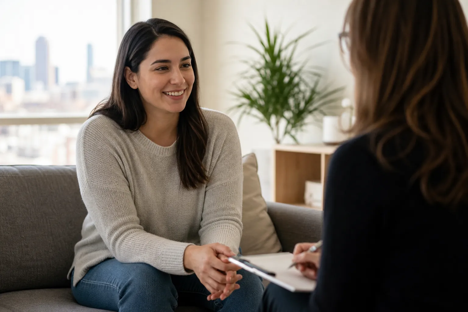 Woman Speaking With a Counselor in a Bright Office A smiling woman sits on a gray sofa facing another woman holding a pen and clipboard in a bright office.