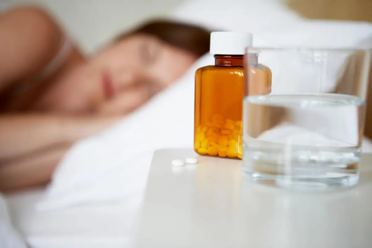 close up of a bottle of pills and a glass of water with sleeping woman in the background asking the question are sleeping pills deadly