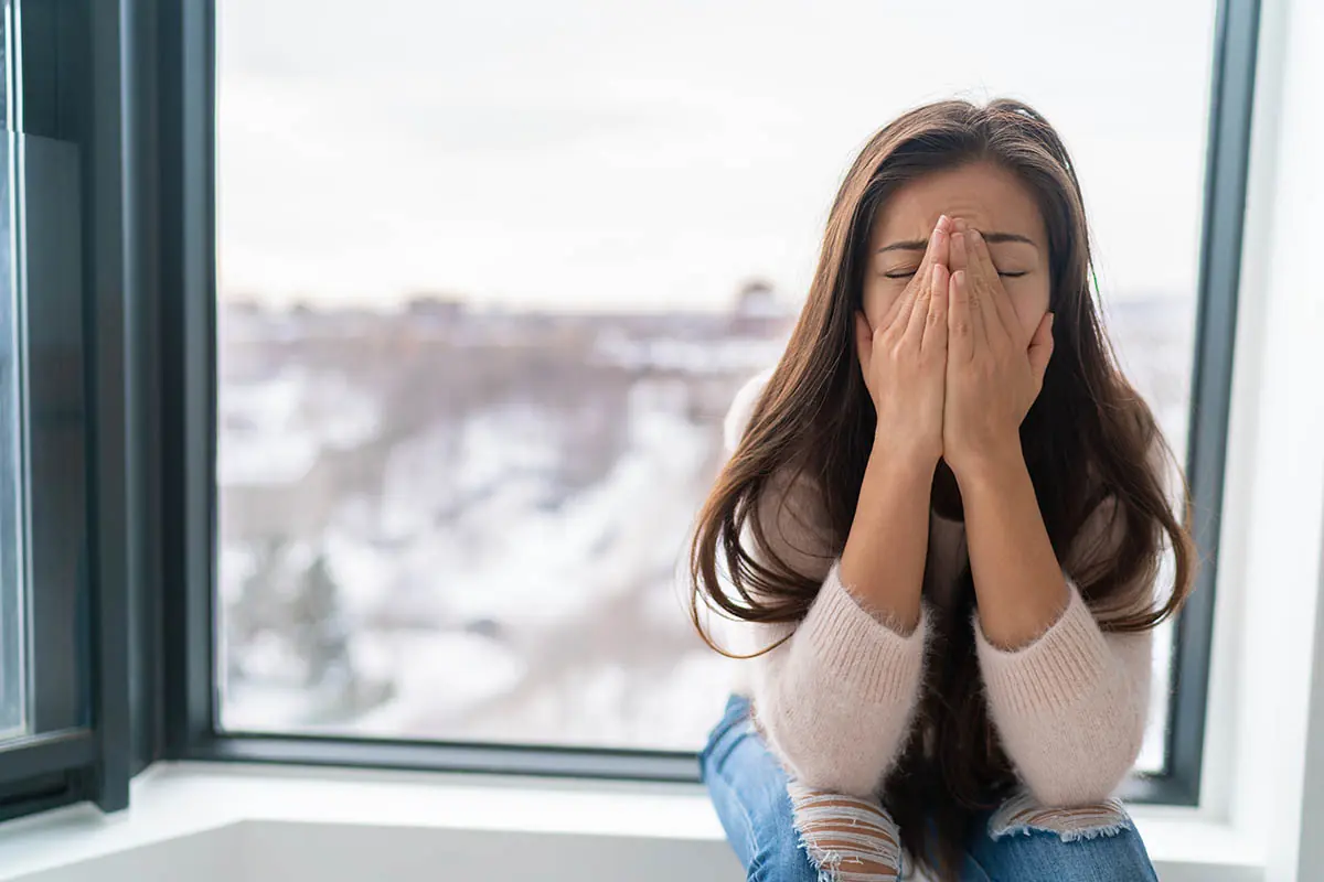 woman sitting on windowsill with a snowy landscape outside and struggling to deal with anxiety in winter