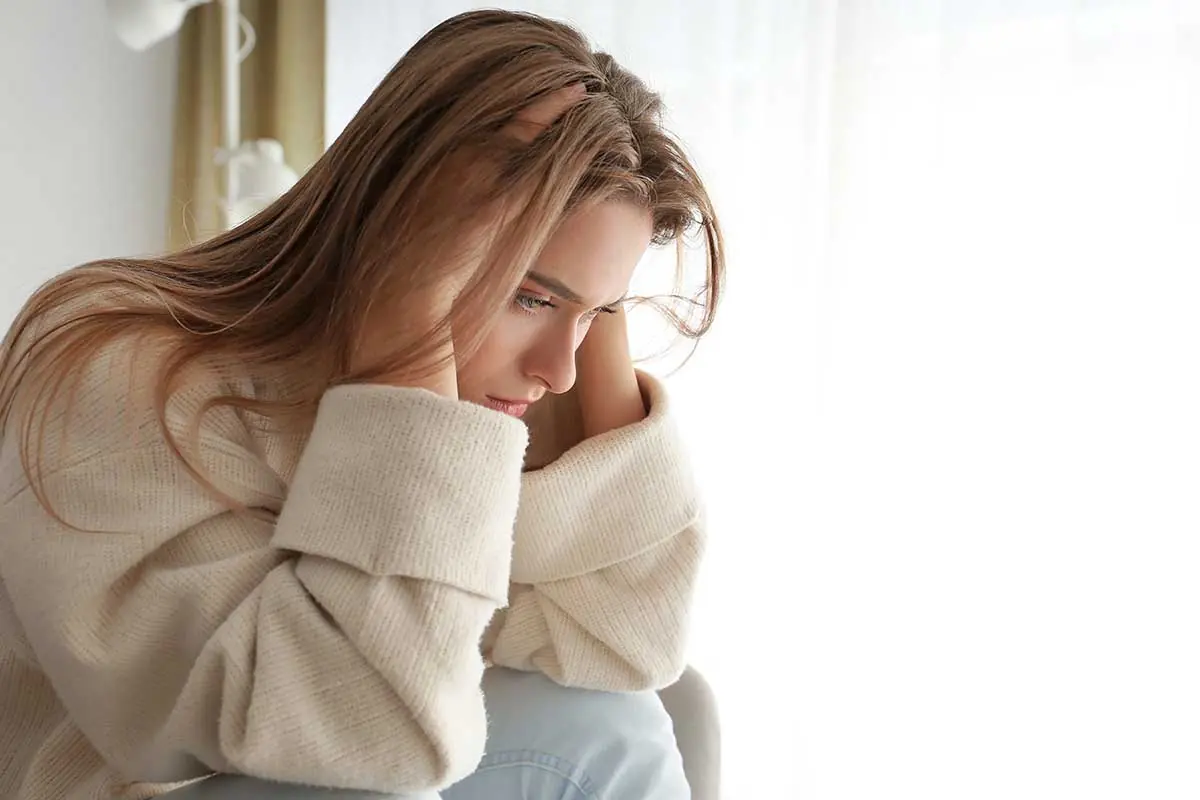 distraught young woman seated by a window with her head in her hands wondering what are the symptoms of depression in women?