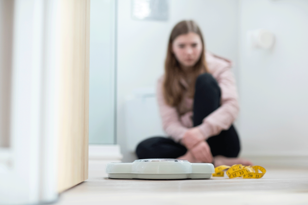 3-Ways-Women-Can-Overcome-Anorexia-Triggers a young woman stares at a scale while struggling to avoid an anorexia trigger