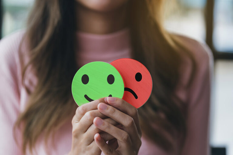 What Are the Bipolar Symptoms in Women_ woman holding up paper cutouts of a green smiley face and red frowning face as a way of asking what are the bipolar symptoms in women