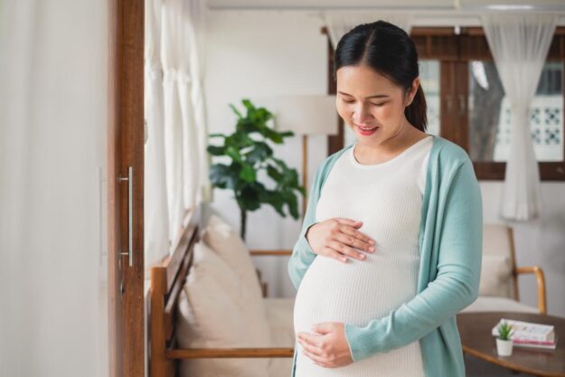 pregnant woman standing in her home holding her belly and pondering safe detox and the fastest way to clean your system during pregnancy.
