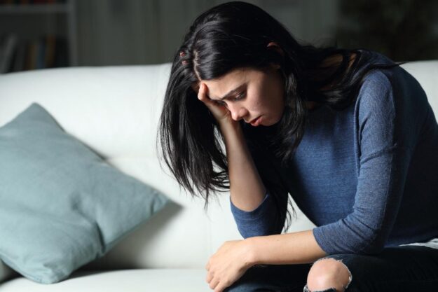 distraught woman seated on her couch at home with her head in her hands not recognizing the signs of borderline personality disorder in womwn like herself.
