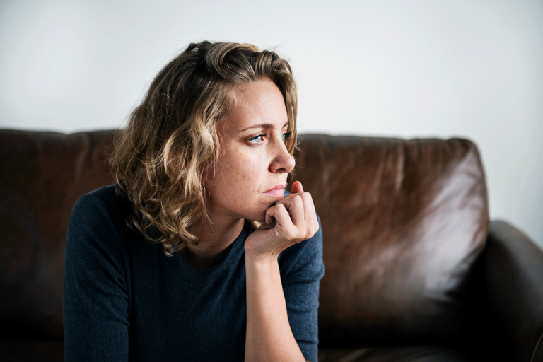 young woman seated on couch deep in thought as she ponders what stops people from getting mental health treatment.