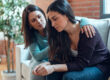 two women seated on a couch together as one consoles the other after spotting common signs of depression in a friend