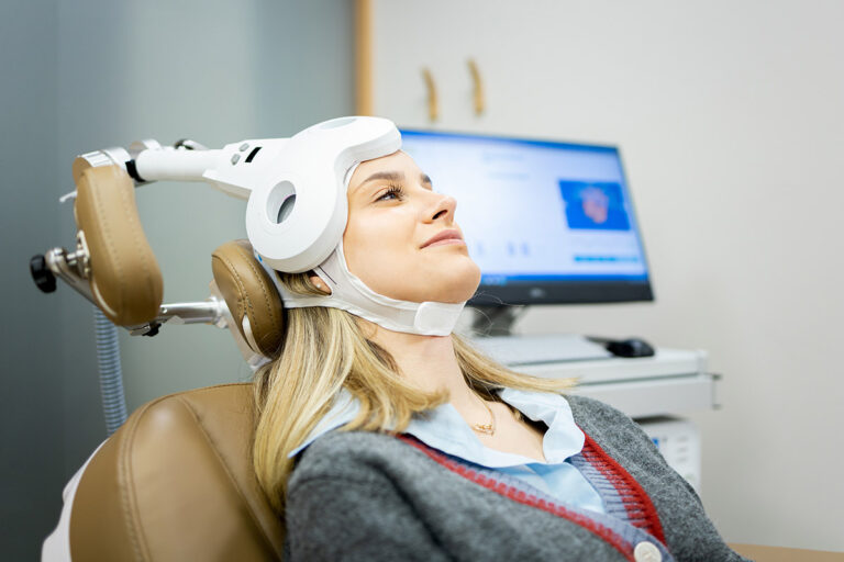A woman sits in a medical chair wearing a transcranial magnetic stimulation (TMS) device on her head, with a computer screen in the background.