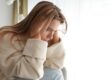 distraught young woman seated by a window with her head in her hands wondering what are the symptoms of depression in women?