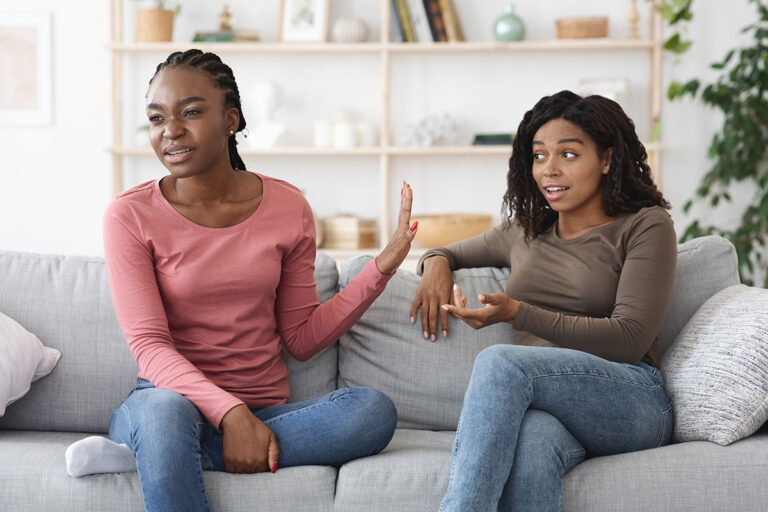 Two sisters seated on couch as one struggles to help her alcoholic sister in denial who is refusing help by offering treatment options