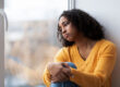 A woman leans against a window. She is experiencing symptoms of PTSD in women.