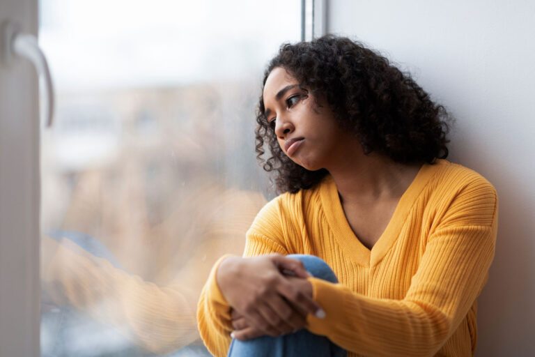 A woman leans against a window. She is experiencing symptoms of PTSD in women.