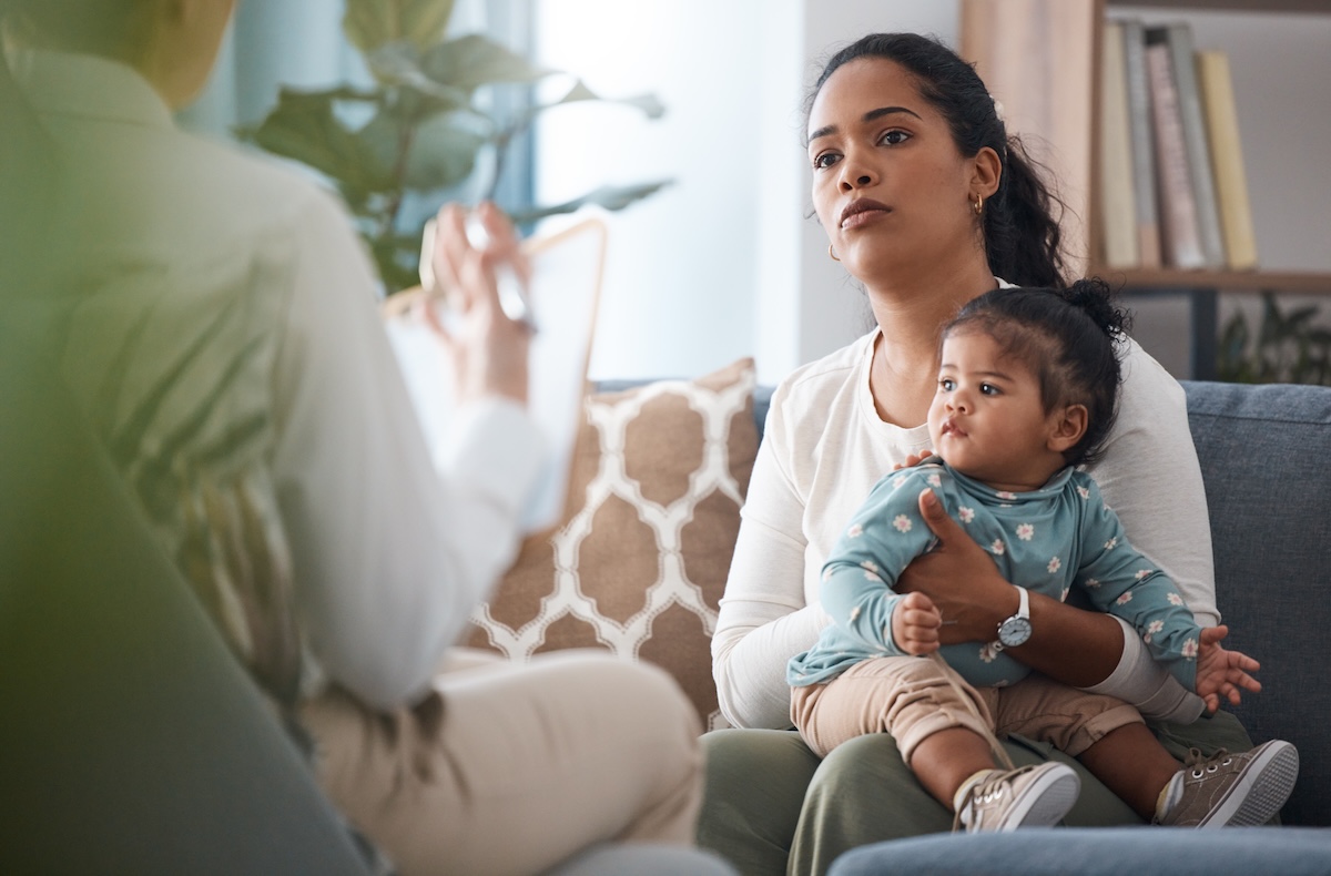 Counseling, therapy and support with baby and mother for postpartum depression, psychology and mental health. Consulting, healthcare and trauma with woman and child in clinic for stress and advice Woman holding a baby on her lap listens attentively to a person speaking and taking notes during a meeting or consultation.