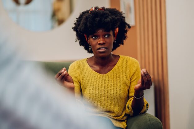 African american woman talking about her mental health on a group therapy session Woman in a yellow sweater gestures with her hands while speaking, appearing engaged in a conversation.